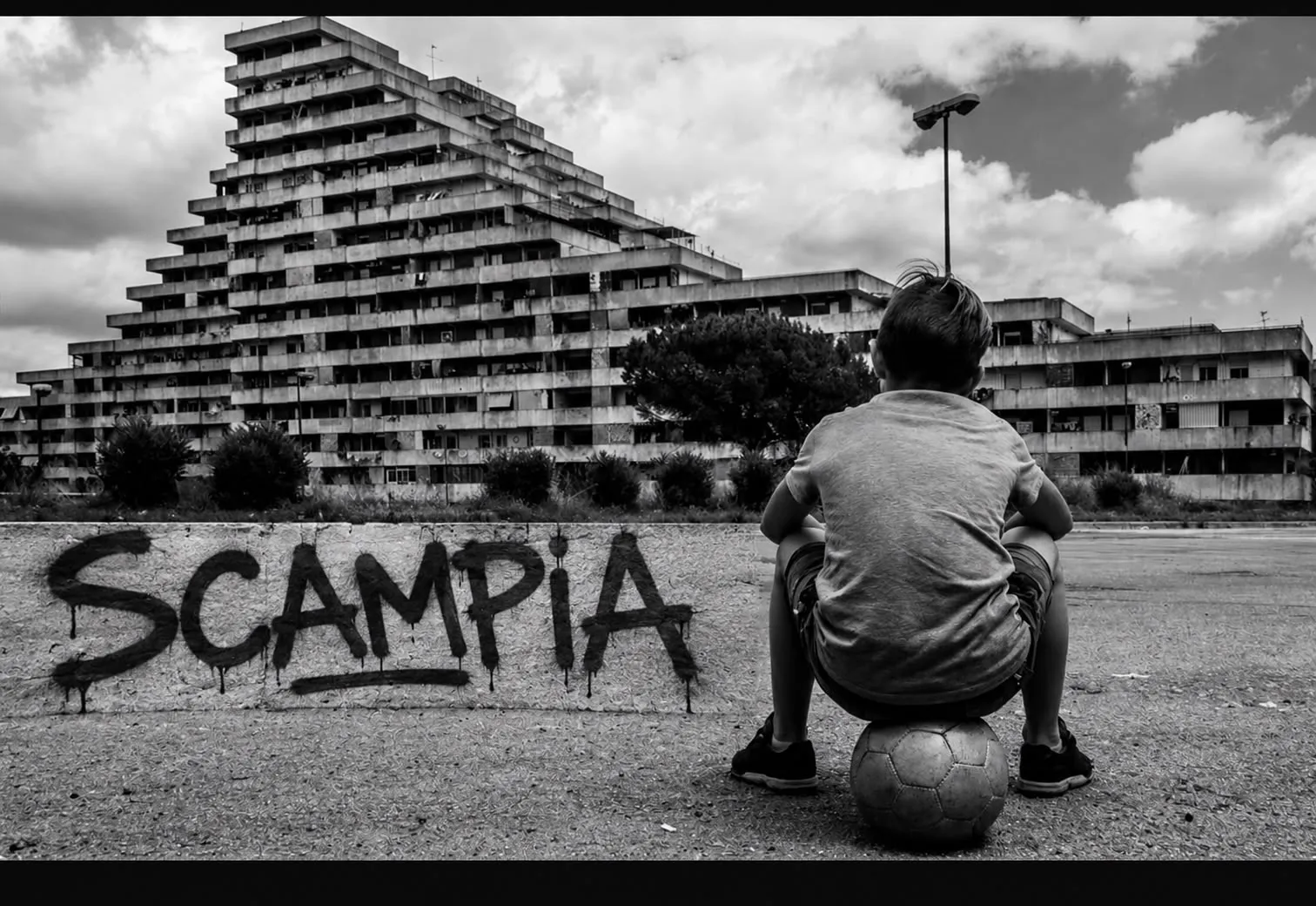 Un bambino seduto su un pallone davanti agli edifici e alla scritta Scampia, in bianco e nero